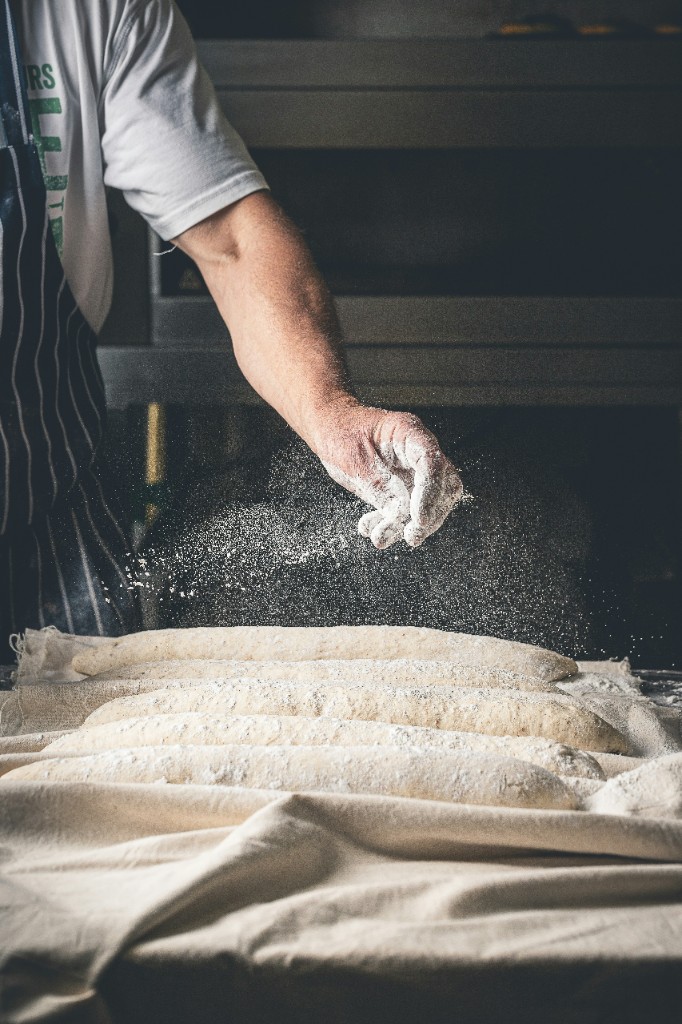 Baker’s hand sprinkling flour over loaves resting on a floured linen couche.